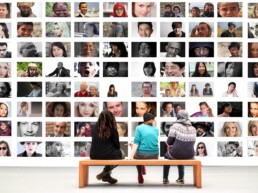 Drei Menschen sitzen auf einer Bank vor einer Wand mit zhalreichen Bilder von Menschen