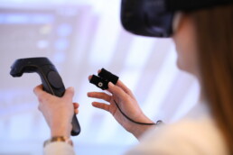 Eine Frau hat eine VR-Brille auf und hält zwei Bedienteile in den Händen.