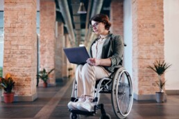 Junge Frau sitzt mit einem Laptop in der Hand im Rollstuhl.