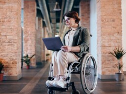 Junge Frau sitzt mit einem Laptop in der Hand im Rollstuhl.