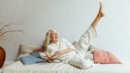 Eine ältere Dame liegt mit einem Buch in der Hand auf dem Sofa, lacht und streckt ein Bein in die Höhe.