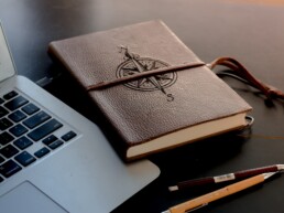 Ein braunes Buch mit eingeprägtem Kompass liegt rechts neben einem Laptop.