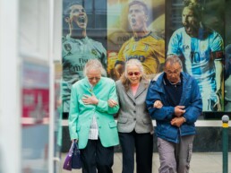 Drei ältere Menschen gehen untergehakt spazieren. Im Hintergrund drei junge berühmte Fußballer auf einem Plakat, deren Kleidungsfarben die Kleidung der Älteren aufgreifen.