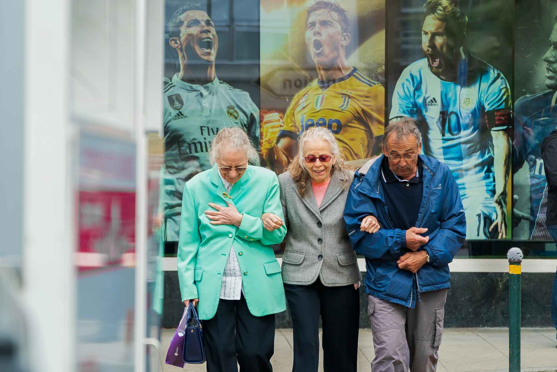Drei ältere Menschen gehen untergehakt spazieren. Im Hintergrund drei junge berühmte Fußballer auf einem Plakat, deren Kleidungsfarben die Kleidung der Älteren aufgreifen.