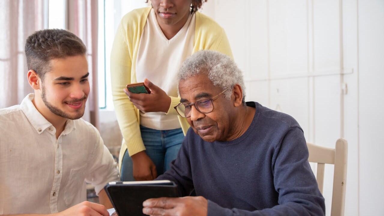 Zwei Pflegepersonen unterstützen einen älteren Herren bei der Nutzung eines Tablets.