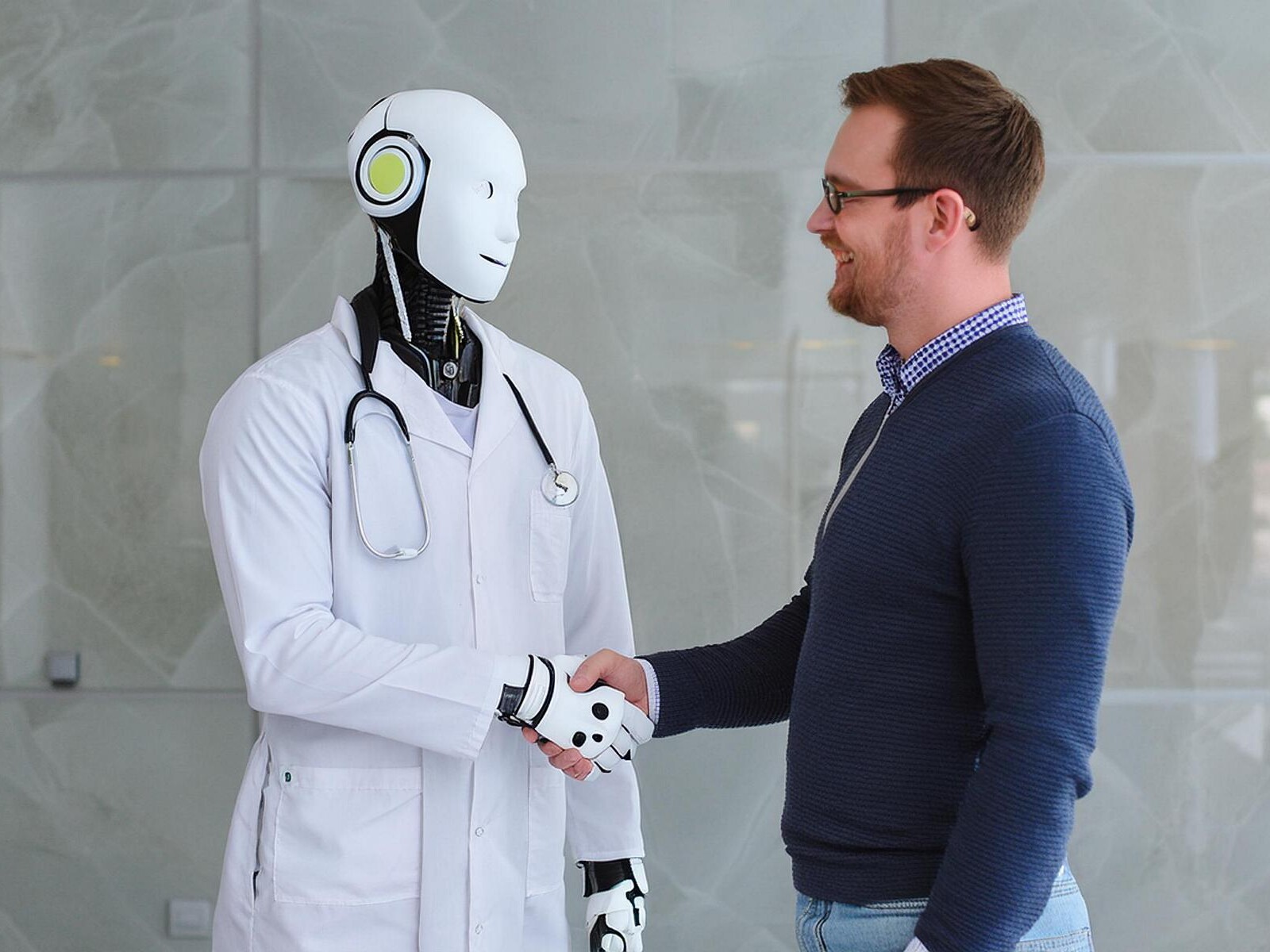 Ein Roboter als Arzt mit Stethoskop um den hals und Arztkittel, er lächelt, rechts neben ihm ein Patient.