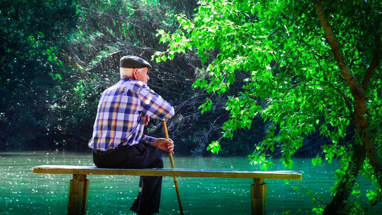 Ein alter Mann mit STock sitzt auf einer Bank an einem See.